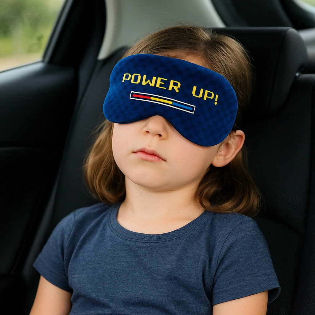 Ein Kind schläft entspannt auf dem Rücksitz eines Autos und trägt eine blaue „Power Up!“ Schlafmaske mit gelber Pixel-Schrift